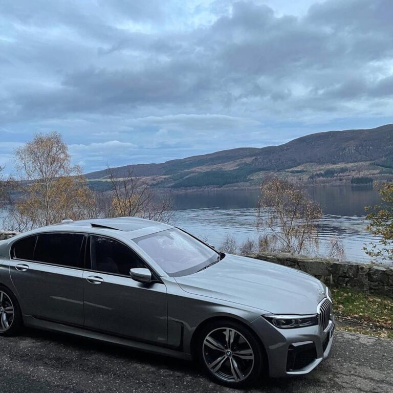 Silver BMW 7 Series parked beside a lake, with hills behind